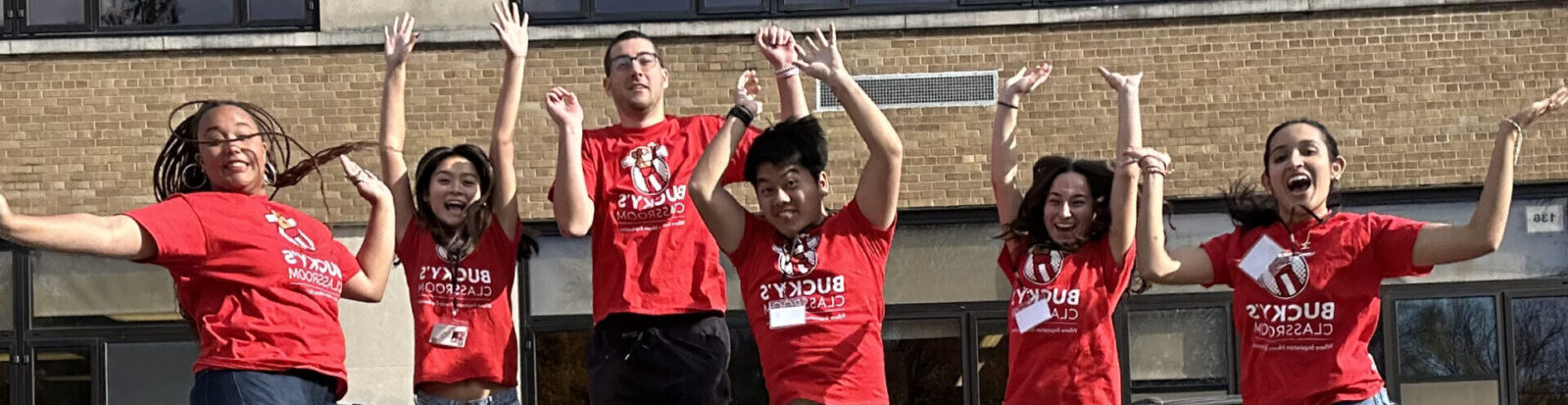 Bucky's Classroom Ambassadors in red t-shorts jumping with their hands raised in front of a school.